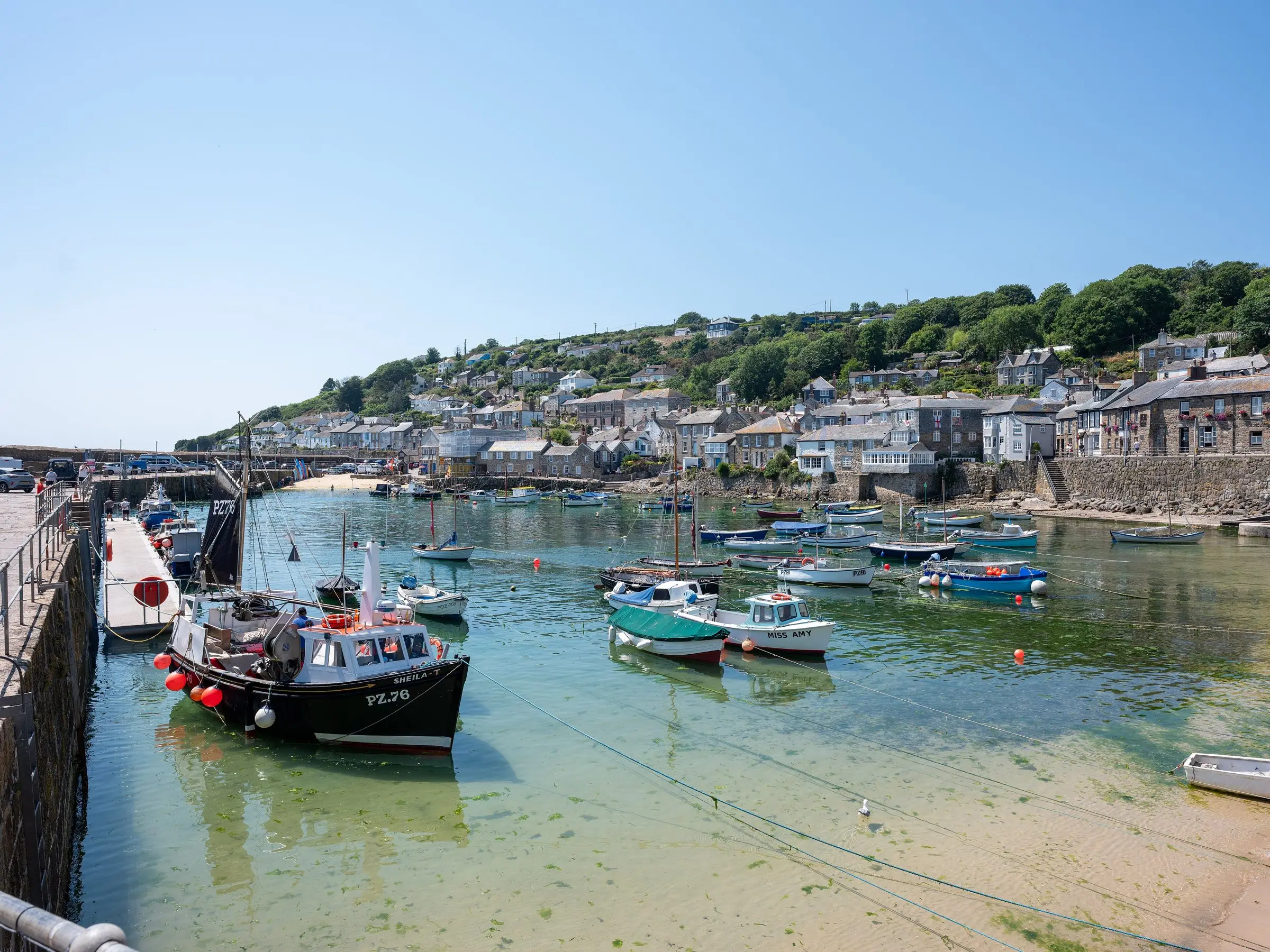 Stone cottages along a Cornish harbour wall on a calm summer morning, small boats moored in clear water