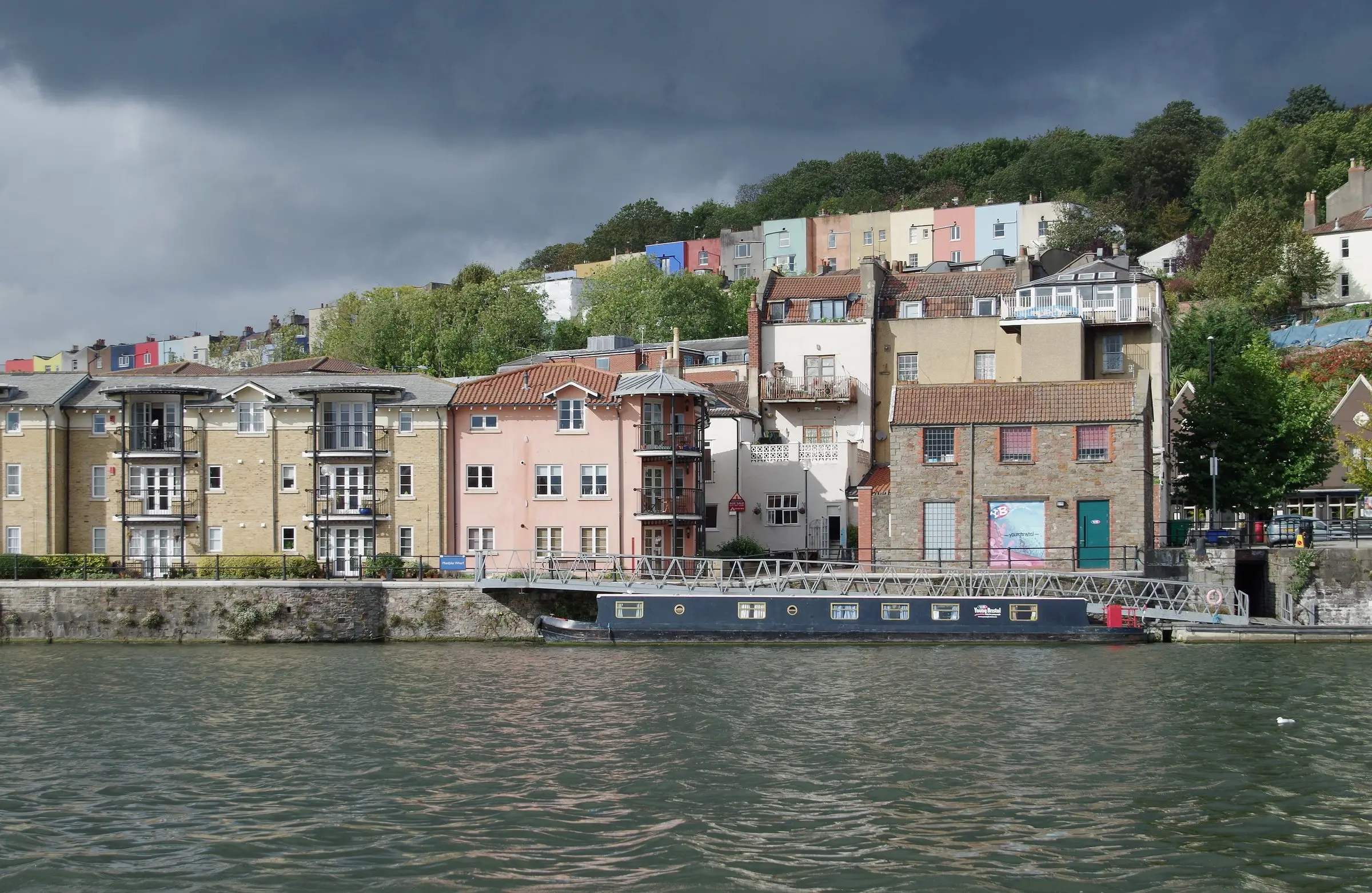 Pastel-painted terraces stacked up the hillside above Bristol's harbour under a dark sky, with a narrowboat moored below