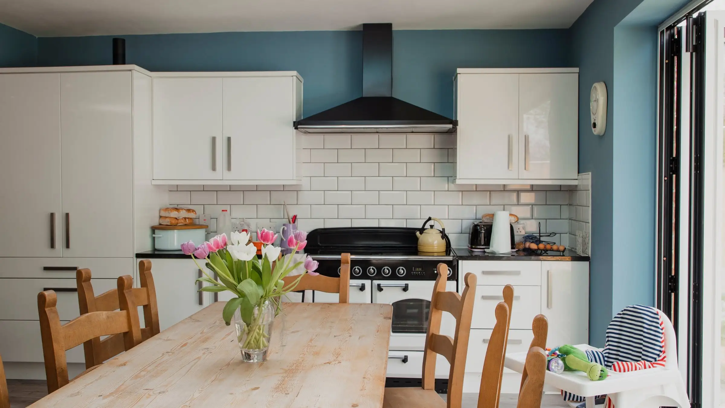 A holiday-let kitchen laid for a family stay: a long wooden table with tulips, a high chair, and garden doors open to the light