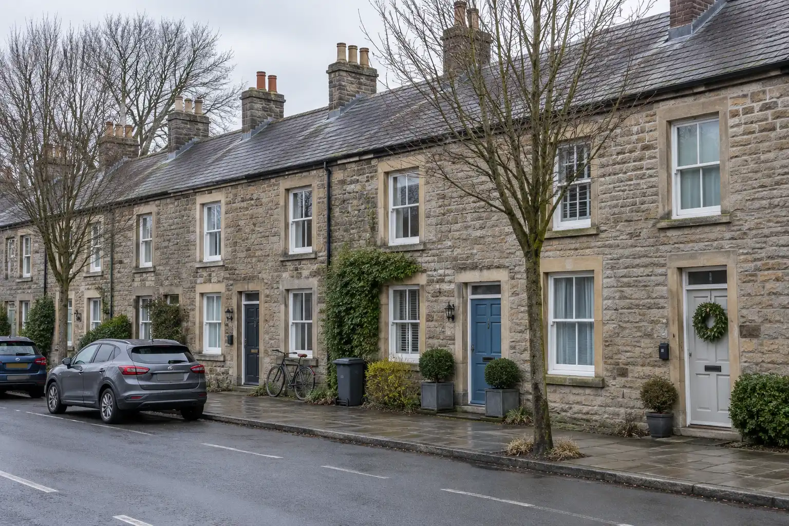 A row of stone terraced cottages on a UK village street in flat winter light, several with key safes beside the doors