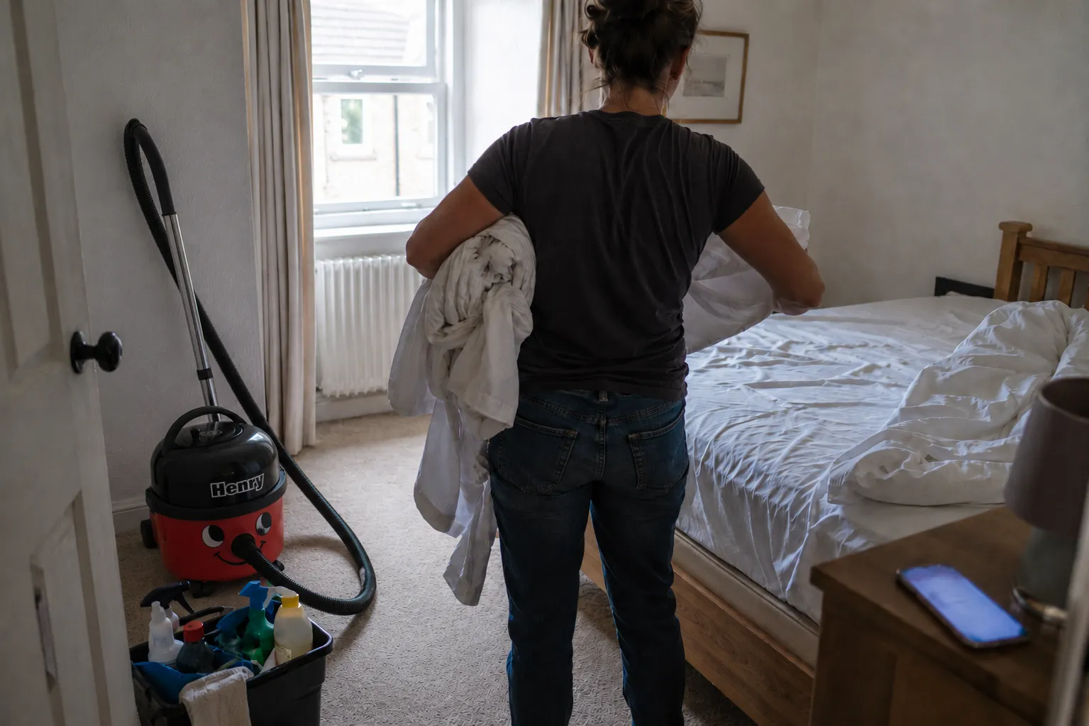 An owner seen from behind stripping a bed in a holiday let, arms full of linen, with a hoover and cleaning caddy waiting